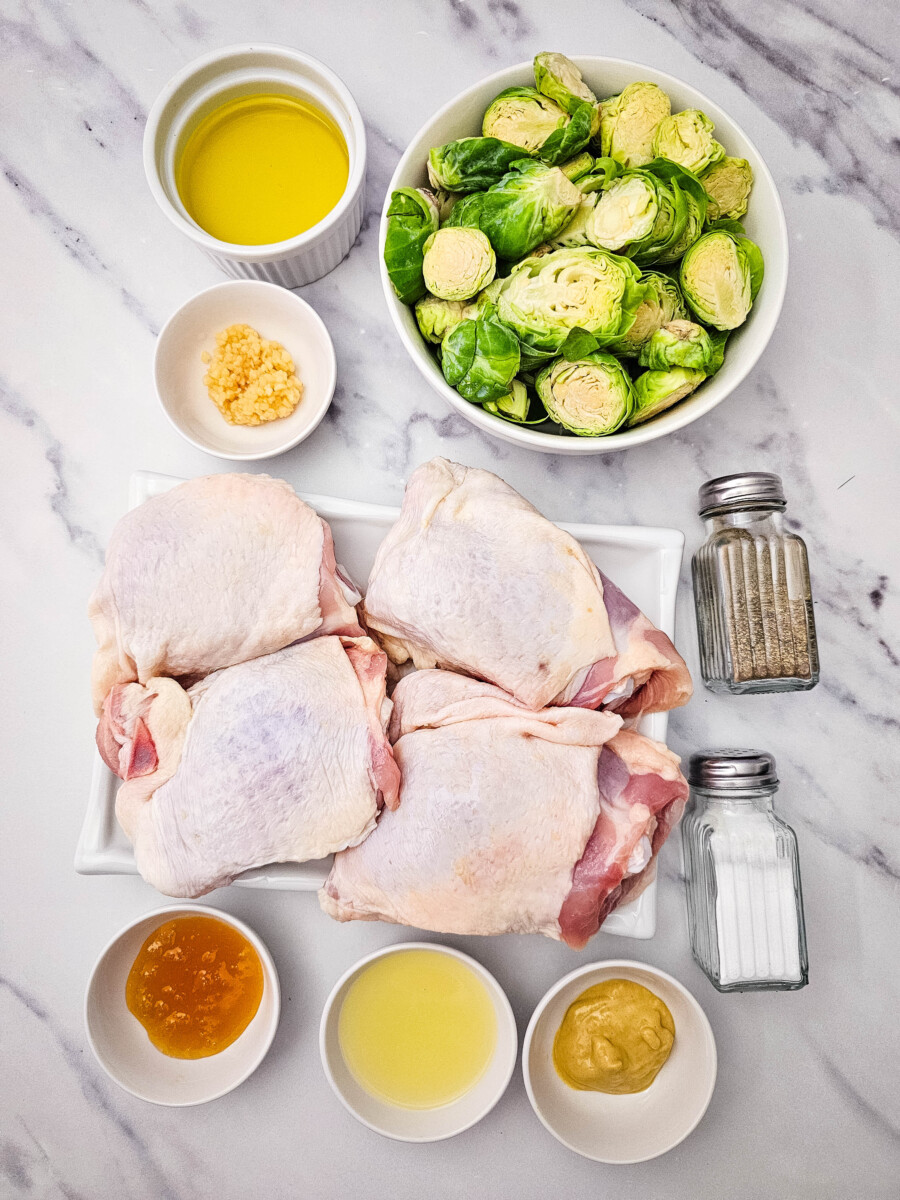 Honey mustard sheet pan chicken thighs ingredients laid out flat including raw chicken thighs, brussels sprouts, olive oil, honey mustard, garlic, and spice jars