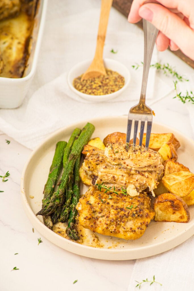 Plated mustard chicken thighs with roasted potatoes and asparagus, fork cutting into seasoned meat