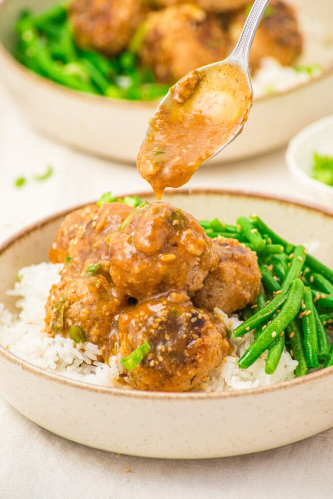Pork meatballs with soy butter sauce served over rice and green beans with sauce being drizzled from a spoon