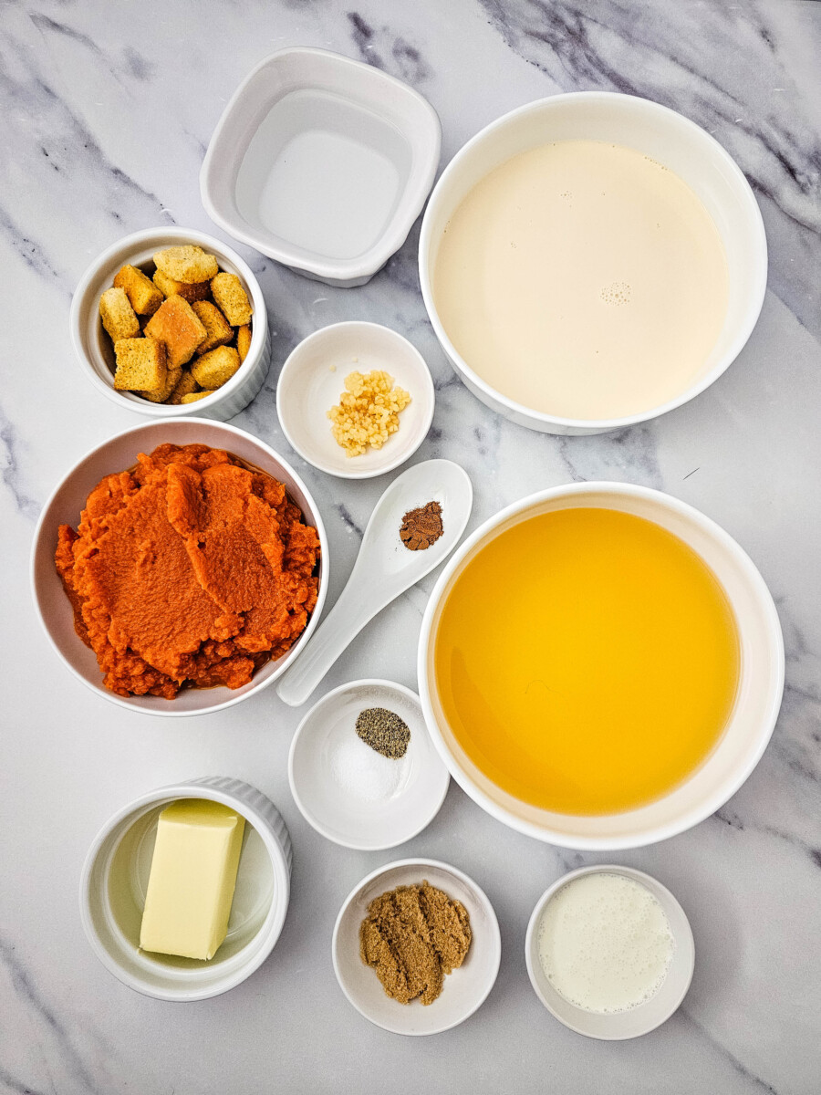 Pumpkin soup ingredients mise en place with pumpkin puree, cream, broth, butter, croutons, and spices arranged in white bowls on marble