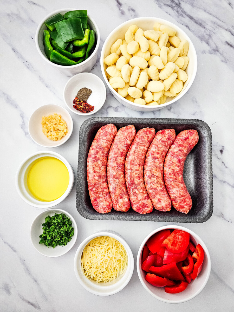 Prepped ingredients for sheet pan sausage and gnocchi laid out overhead including sausages, gnocchi, vegetables, cheese, and seasonings