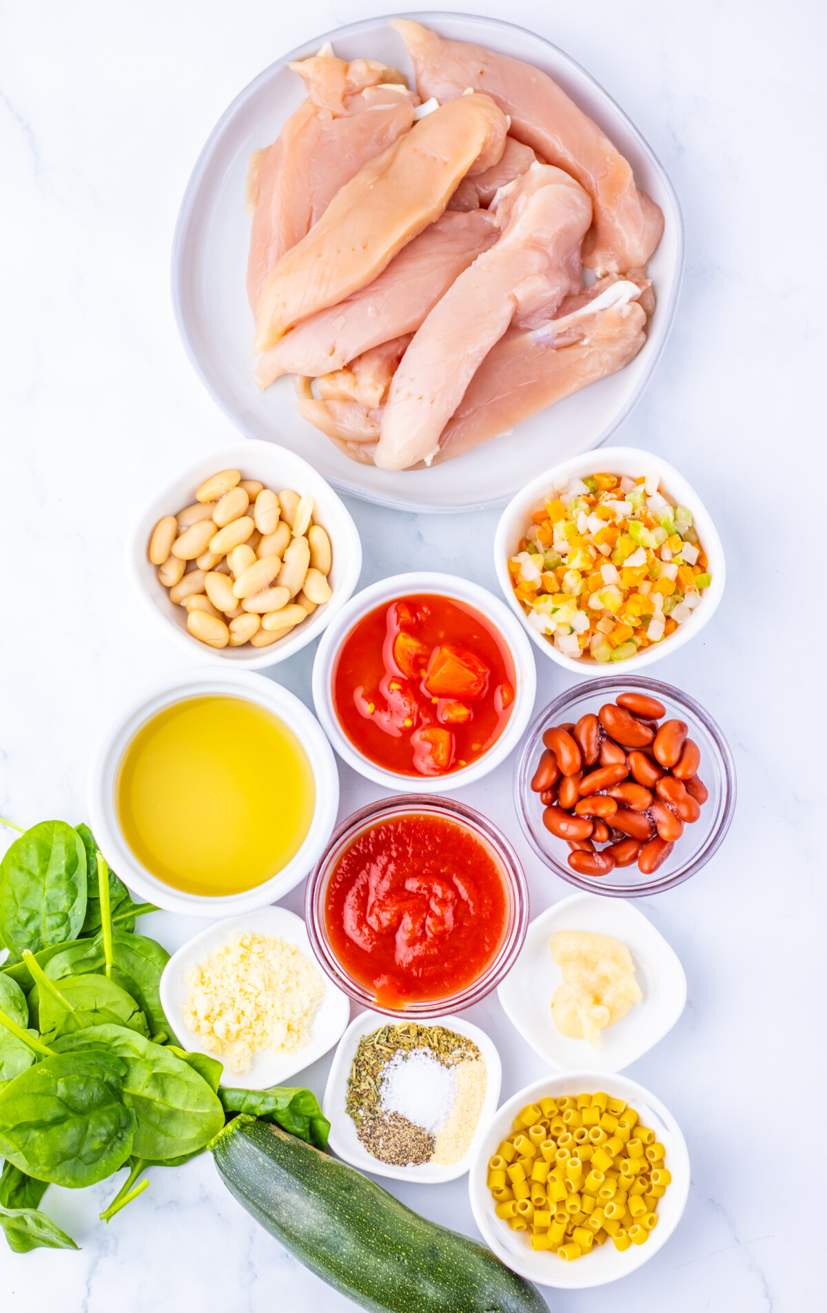 Slow cooker minestrone soup ingredients laid out including raw chicken, beans, corn, diced tomatoes, spinach, zucchini, and seasonings in white bowls