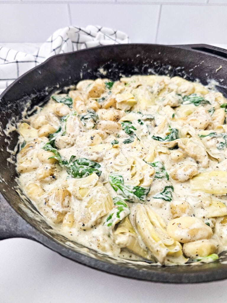 Spinach artichoke gnocchi cooking in skillet with creamy sauce and fresh herbs