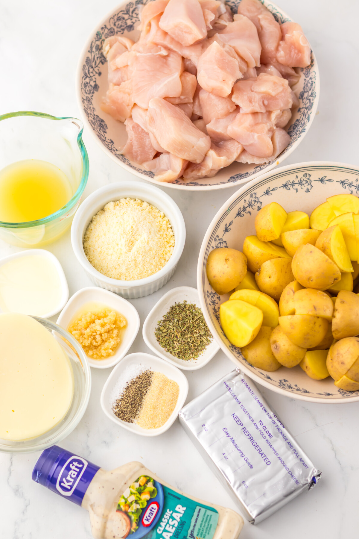 Ingredients for slow cooker cheesy Caesar chicken and potatoes including raw chicken, potatoes, cream, and seasonings arranged on marble counter