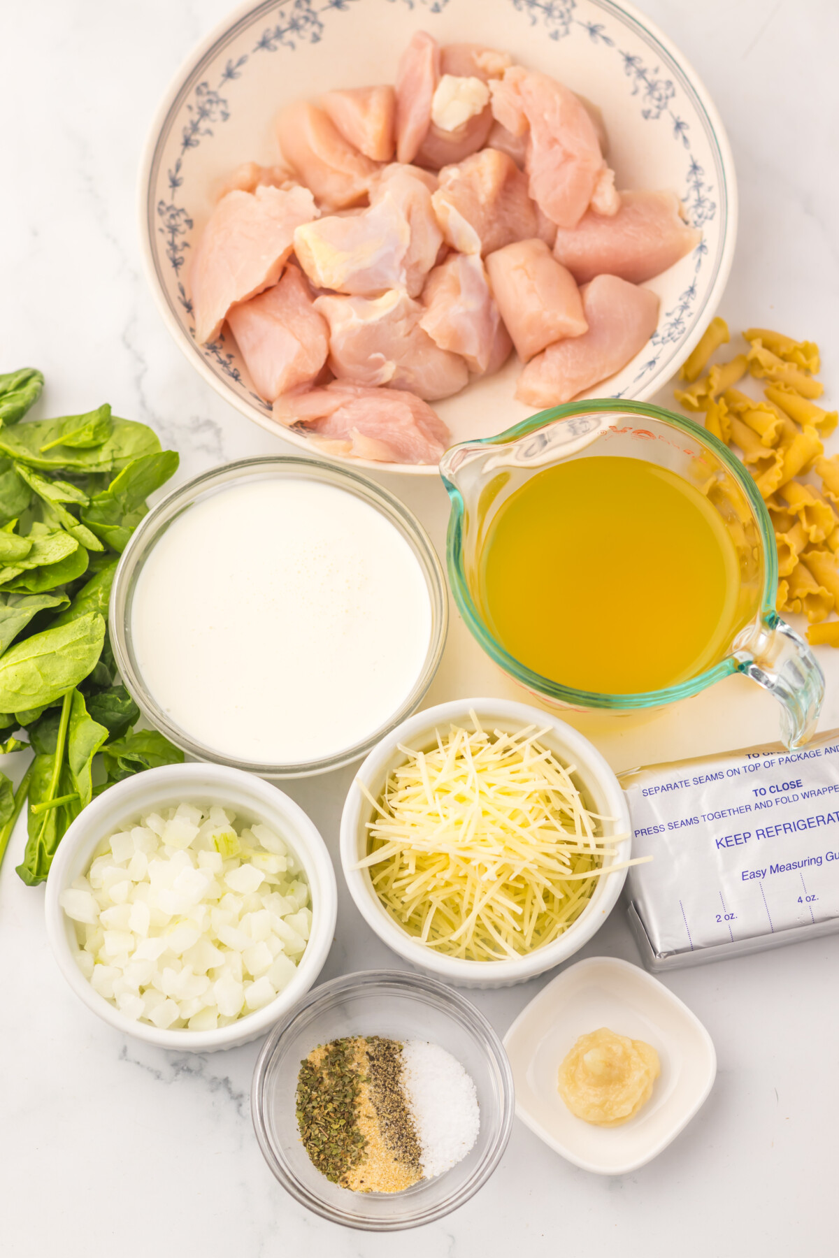 Slow cooker chicken alfredo soup ingredients laid out including raw chicken, cream, oil, pasta, cheese, onions, garlic and seasonings