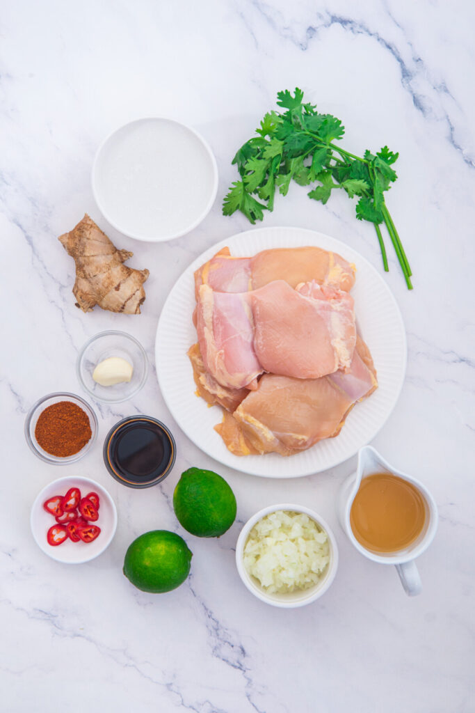 Coconut Lime Chicken ingredients laid out flat including raw chicken breasts, lime, fresh cilantro, ginger, garlic, and Asian seasonings on marble background