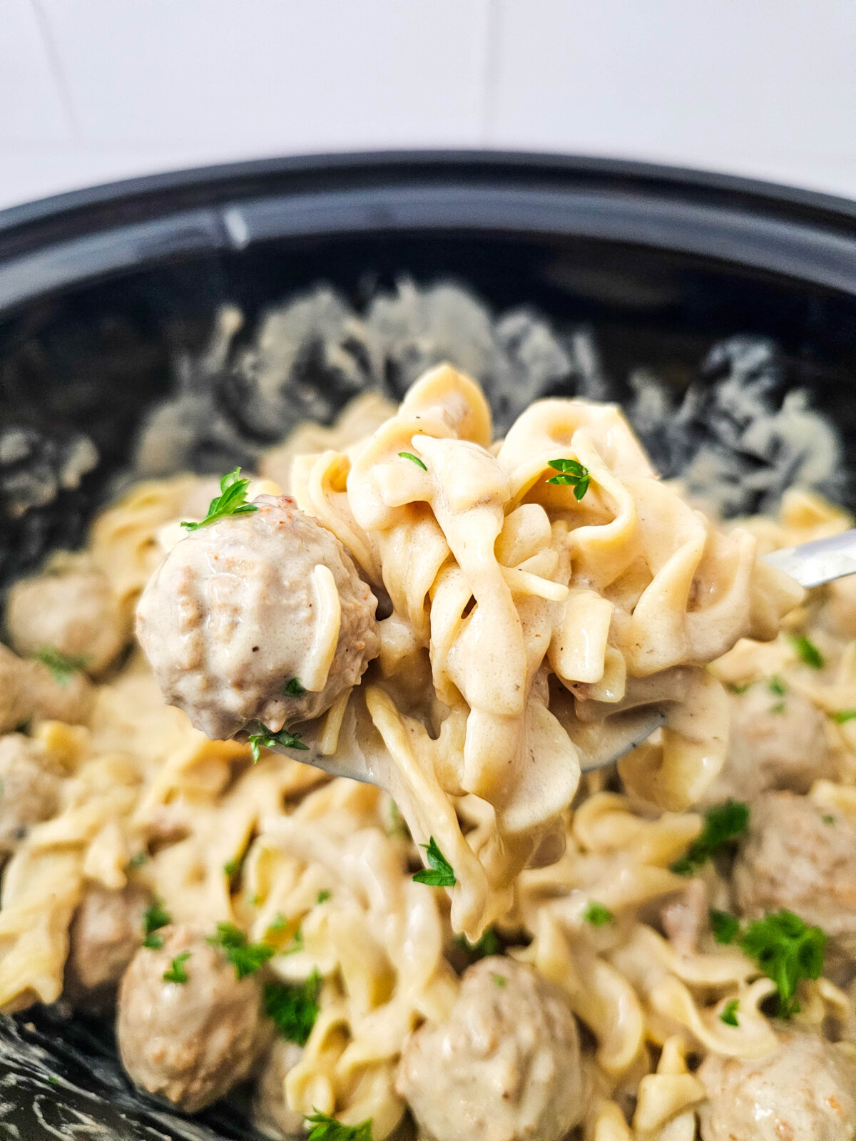 Crockpot Swedish meatballs with egg noodles in creamy sauce being served with a spoon, garnished with parsley