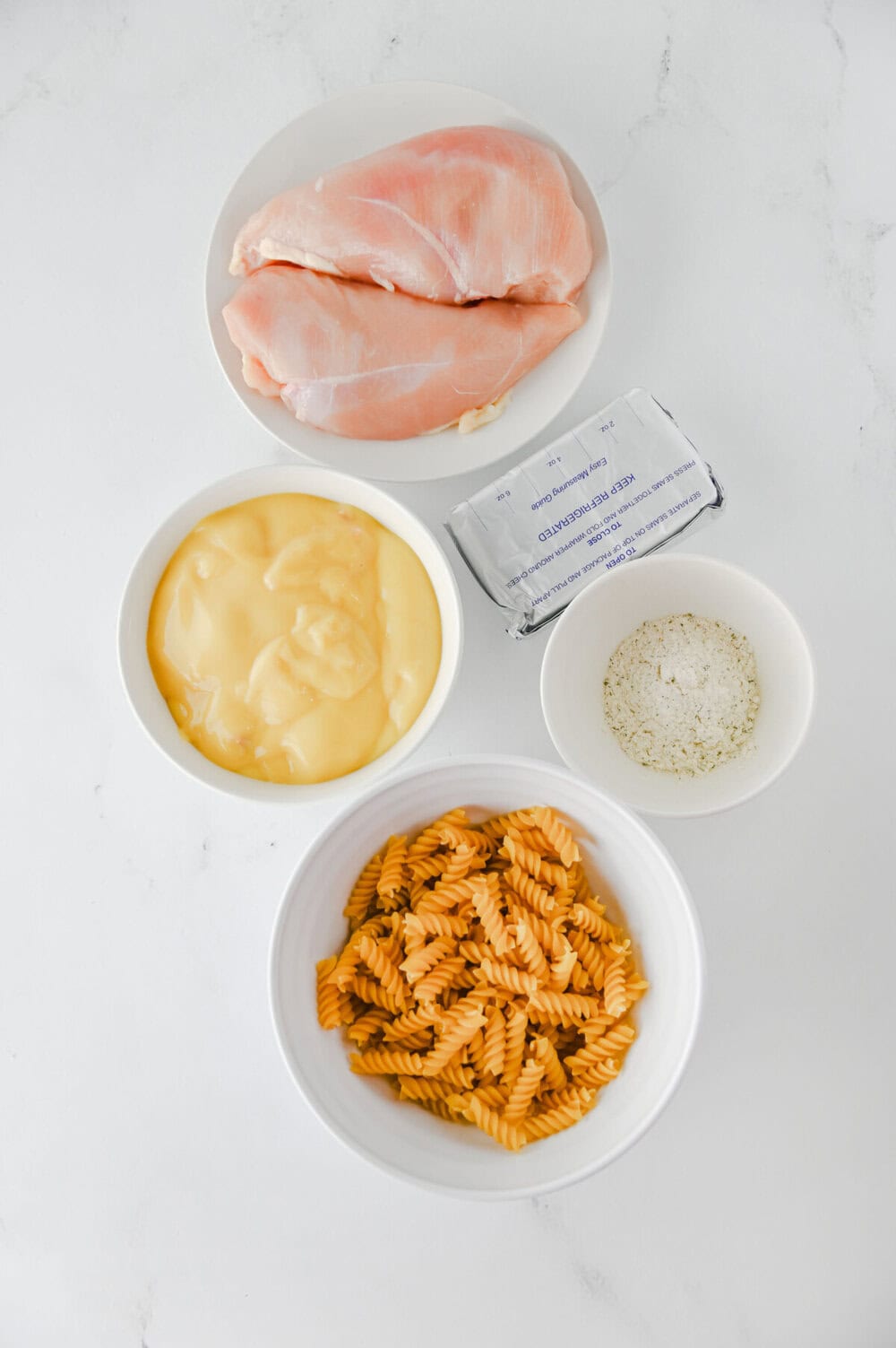 Crockpot ranch chicken pasta ingredients laid out in bowls: raw chicken breasts, mayo, breadcrumbs, rotini pasta, and butter