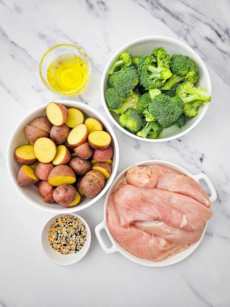 Sheet pan chicken ingredients laid out in bowls: potatoes, broccoli, chicken breast, olive oil, and everything bagel seasoning on marble countertop