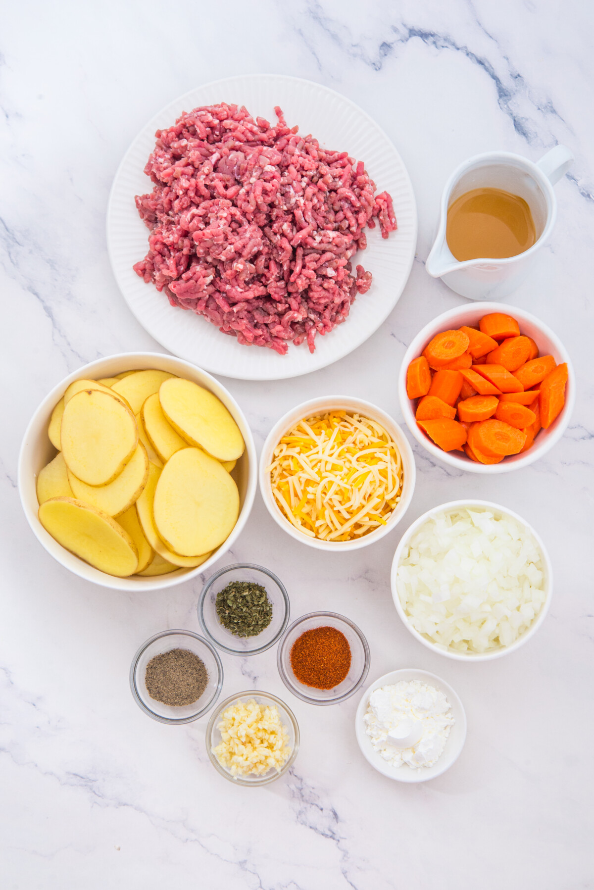 Slow cooker beef and potatoes ingredients including ground beef, sliced potatoes, carrots, cheese, onions, and seasonings in white bowls