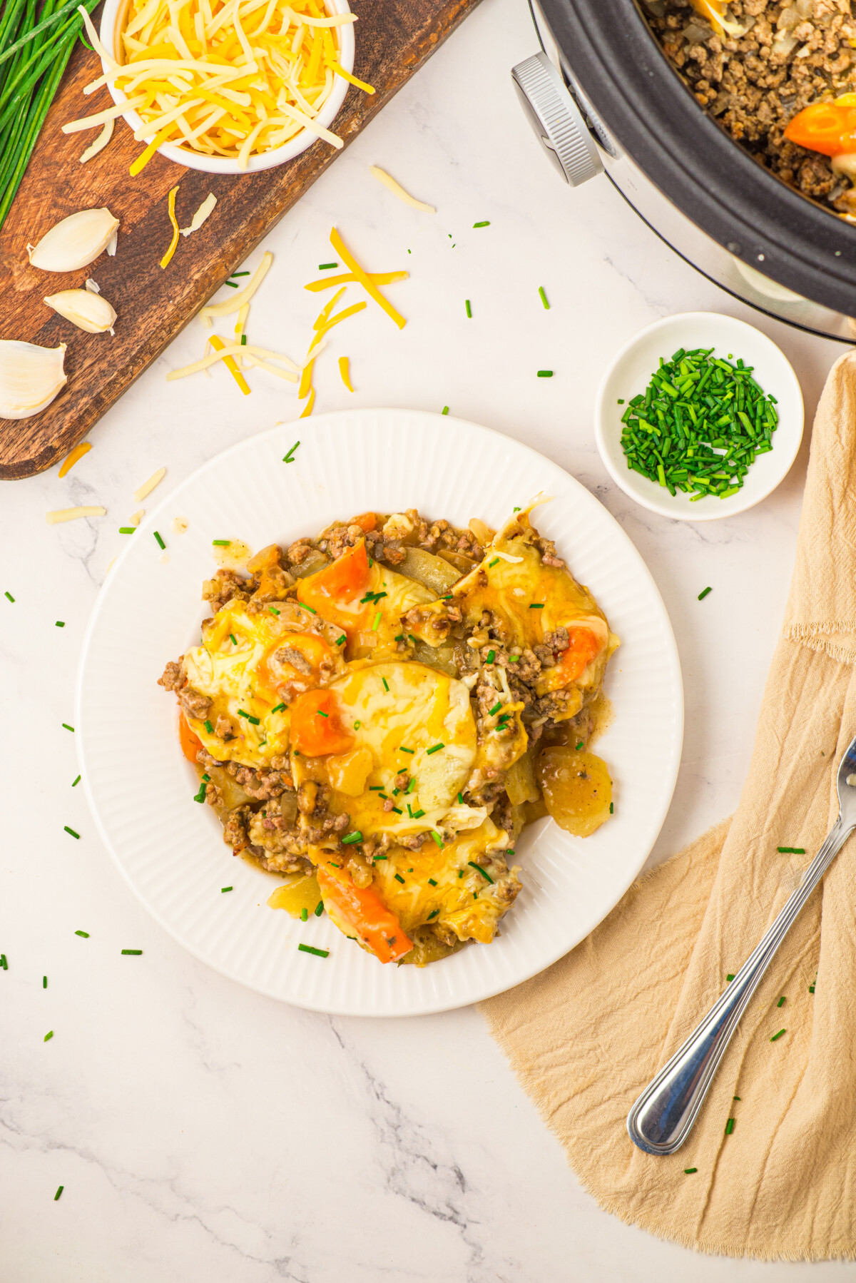 Slow cooker layered beef and potatoes with melted cheddar cheese and chive garnish on white plate
