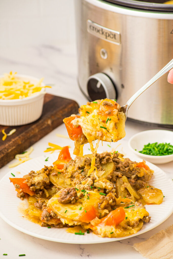 Slow cooker beef and potatoes casserole on white plate with spoon lifting a portion, showing ground beef, diced potatoes and peppers garnished with chives